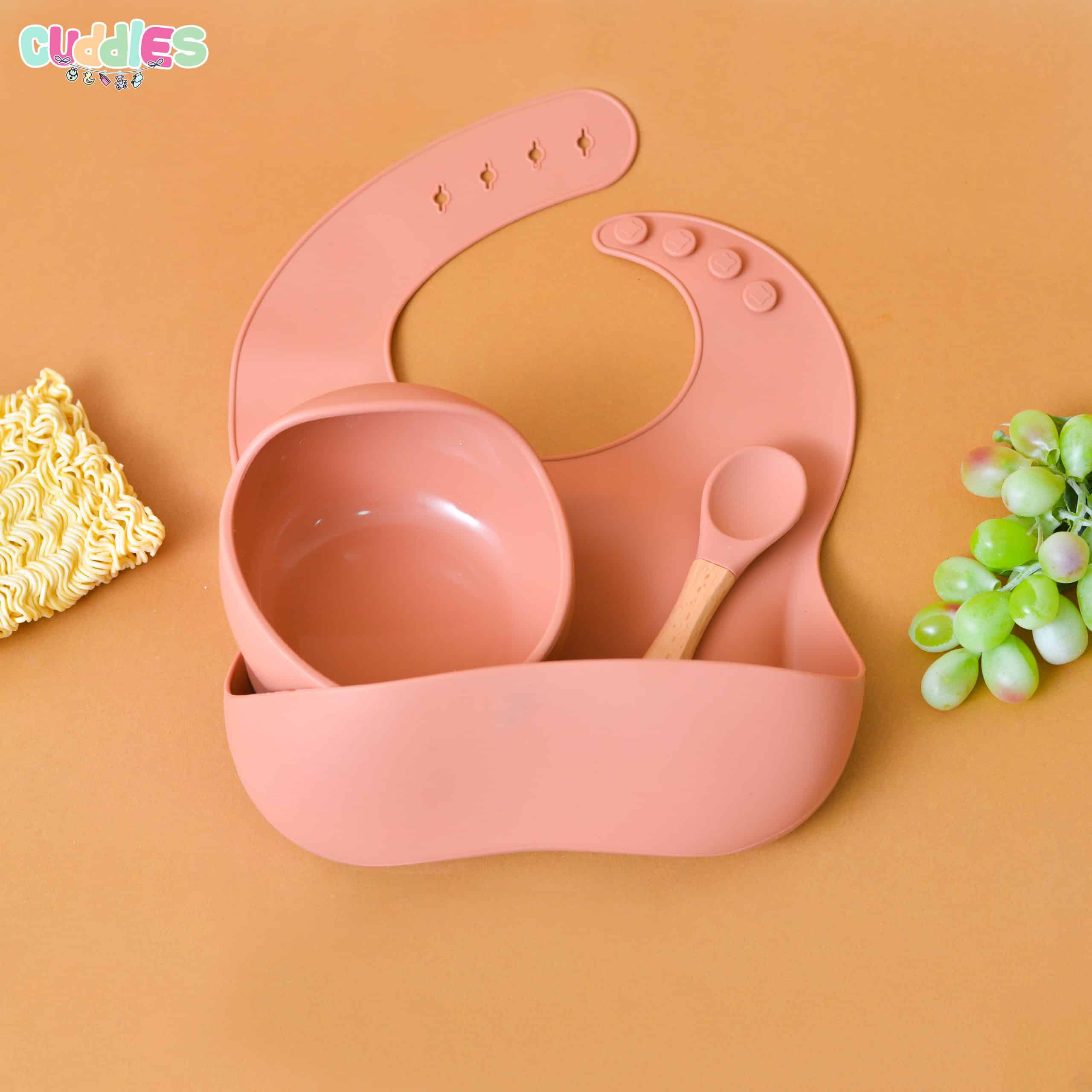 Colorful silicone bib set displayed with a matching suction bowl and easy-grip spoon, all made from BPA-free material. The bib has an adjustable neck strap and a deep front pocket to catch food spills, and the entire set is shown on a clean, neutral background highlighting its mess-free, child-friendly design.